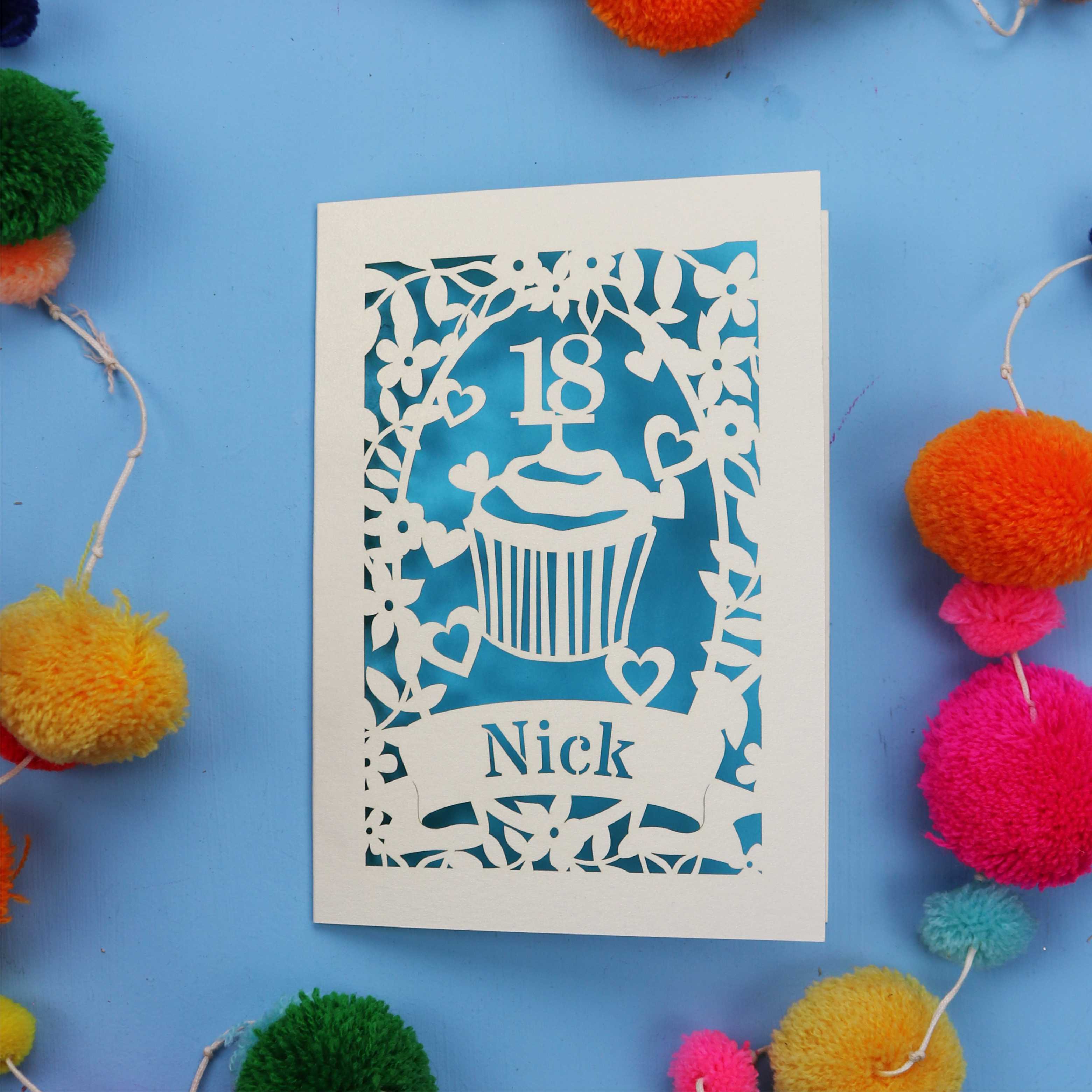 Decorative 18th birthday card with cut-out design on a blue background with colourful pom-poms.