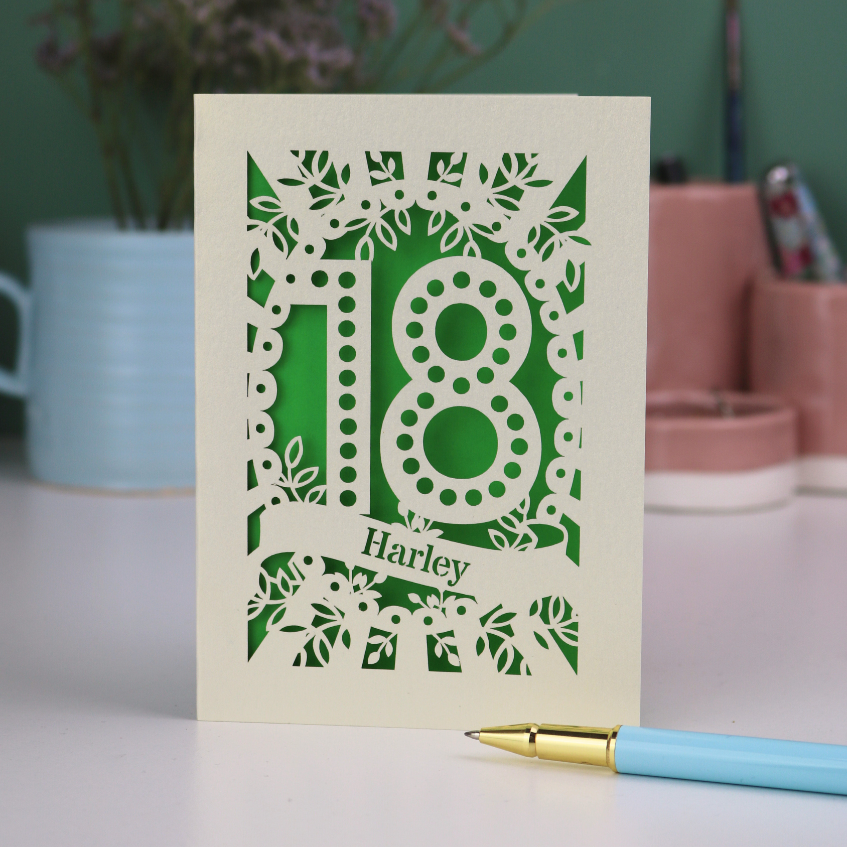Personalised 18th birthday Card with laser-cut number 18 and name 'Harley' on a green background, placed on a surface with a pen and decorative items in the background.