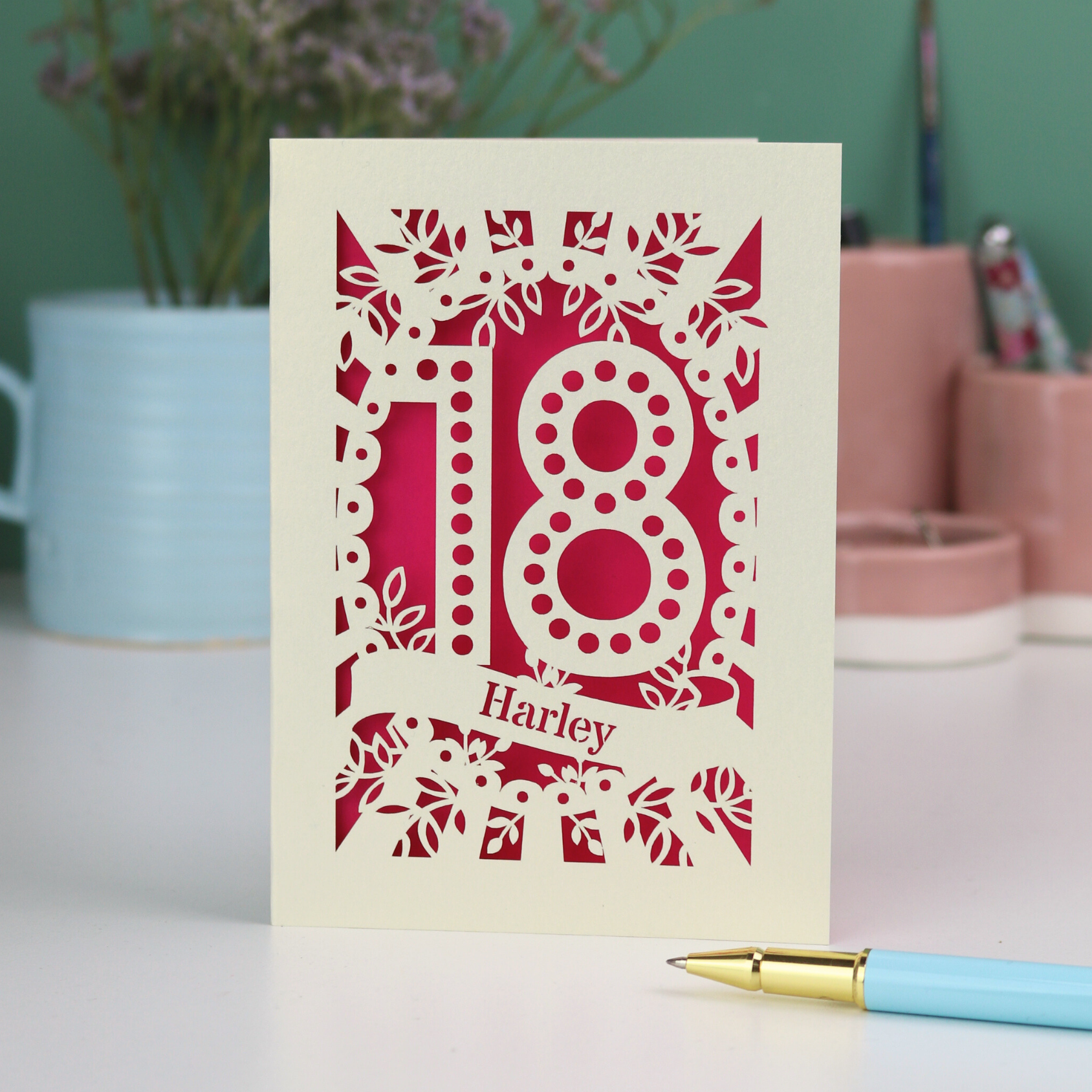 Personalised 18th birthday Card with laser-cut number 18 and name 'Harley' on a shocking pink background, placed on a surface with a pen and decorative items in the background.