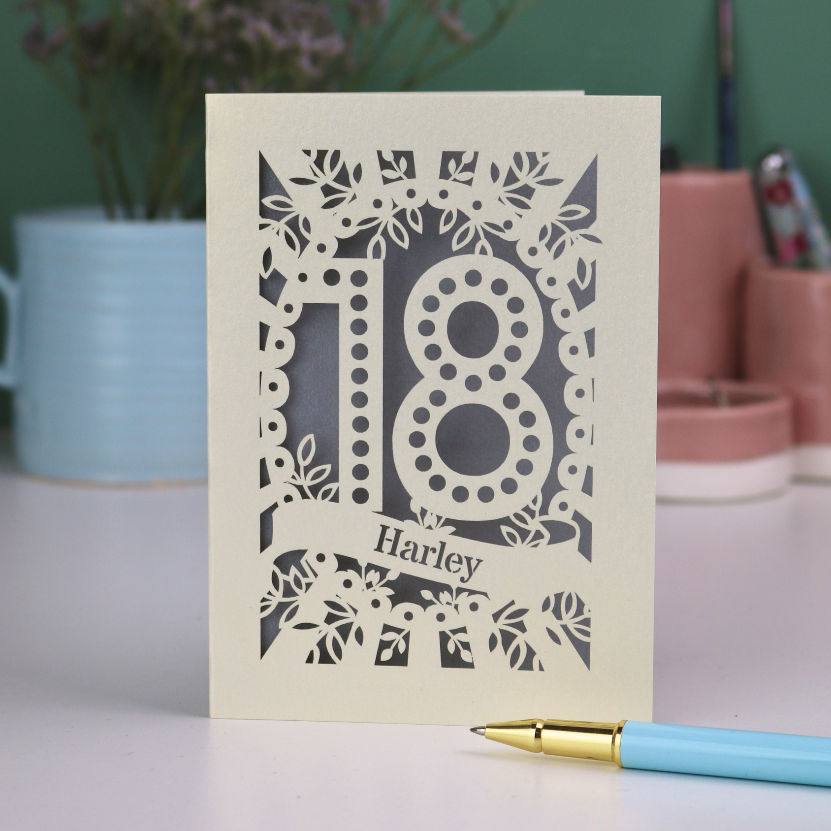 18th Birthday Card with laser-cut number 18 and personalized name, placed on a surface with a pen and decorative items in the background.