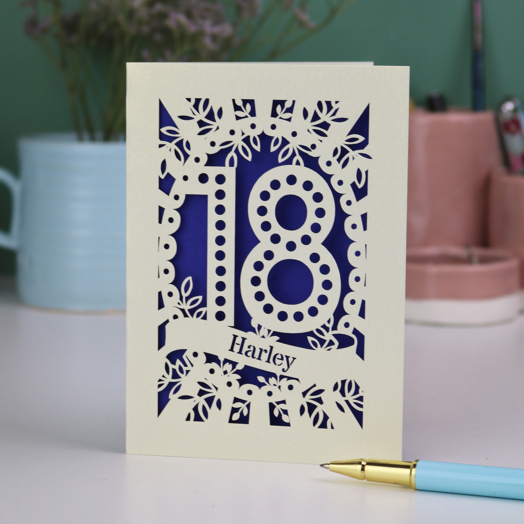 Personalised 18th birthday Card with laser-cut number 18 and name 'Harley' on a green background, placed on a surface with a pen and decorative items in the background.