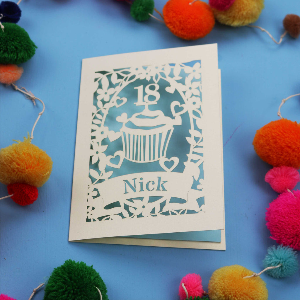 Decorative 18th birthday card with cut-out design on a blue background with colourful pom-poms.