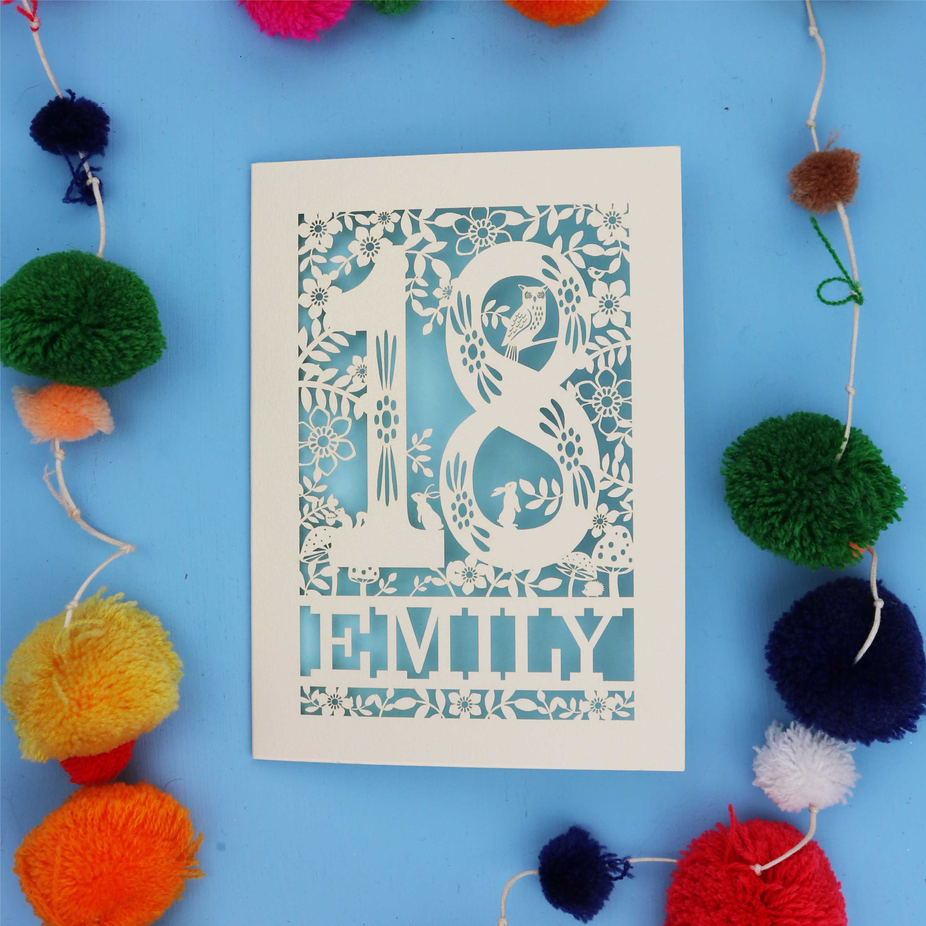 Special 18th birthday card with floral patterns and 'EMILY' text on a blue background with colourful pom-poms.