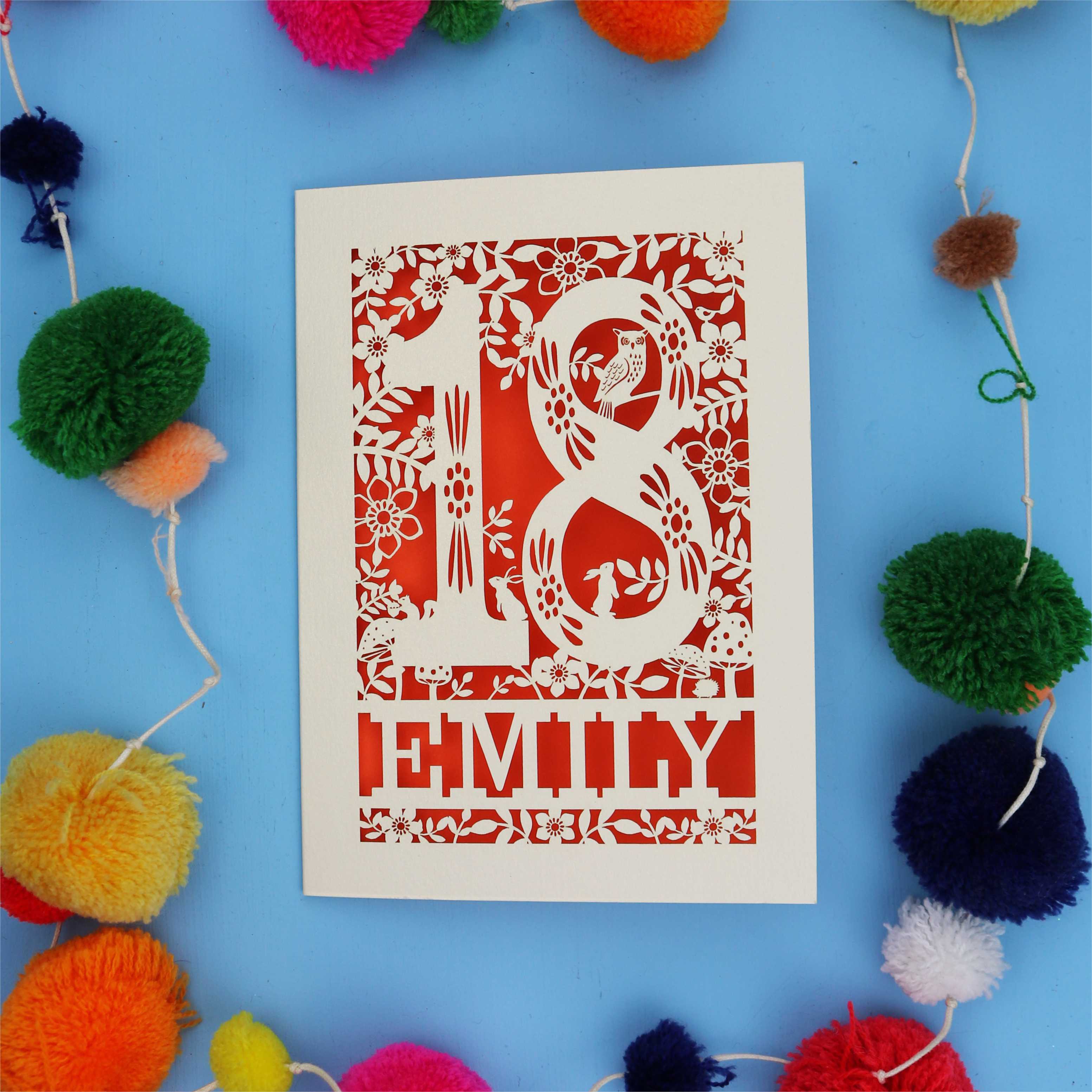 Decorative paper cut 18th birthday card  with floral patterns and 'EMILY' text on a blue background with colourful pom-poms.