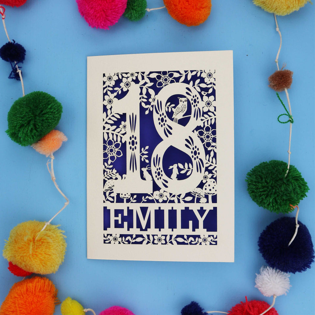 Decorative paper cut 18th birthday card  with floral patterns and 'EMILY' text on a blue background with colourful pom-poms.