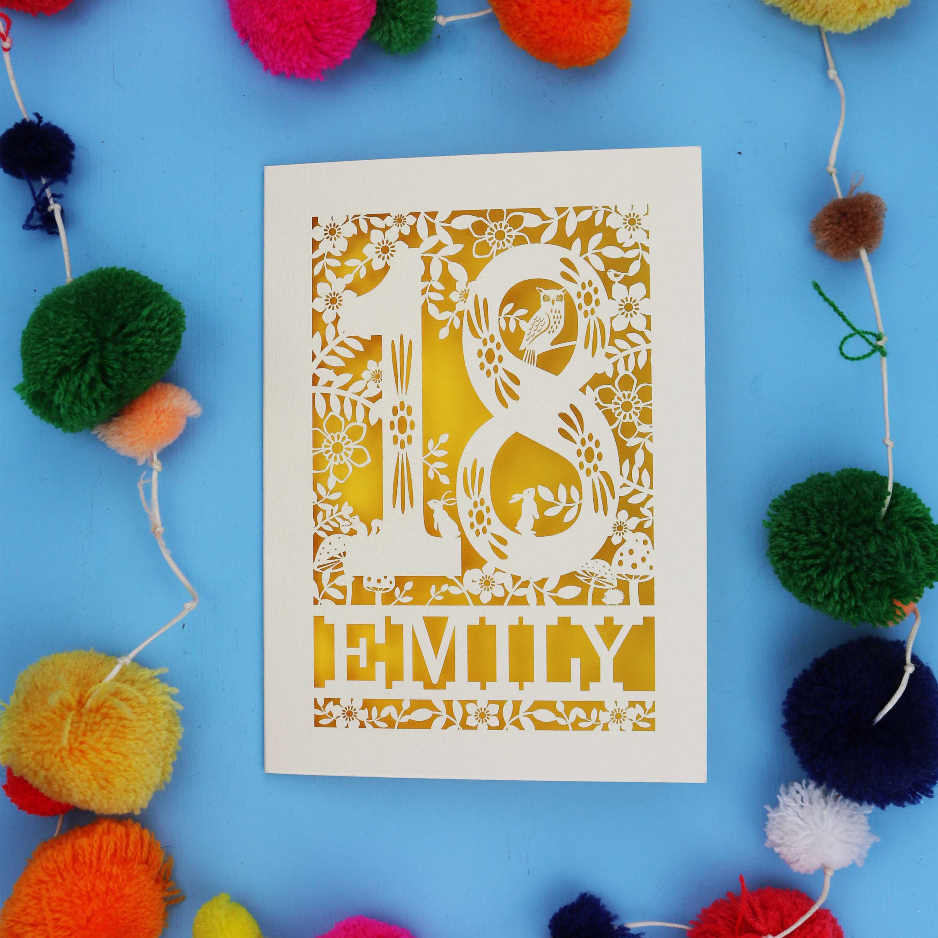 Pretty 18th birthday card with floral patterns and 'EMILY' text on a blue background with colourful pom-poms.