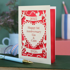 A laser cut cream first anniversary card with banners and heart design and the text "Happy 1st Anniversary Lia & Cam" with a banner above and the year 2025, and a banner below with the year 2026