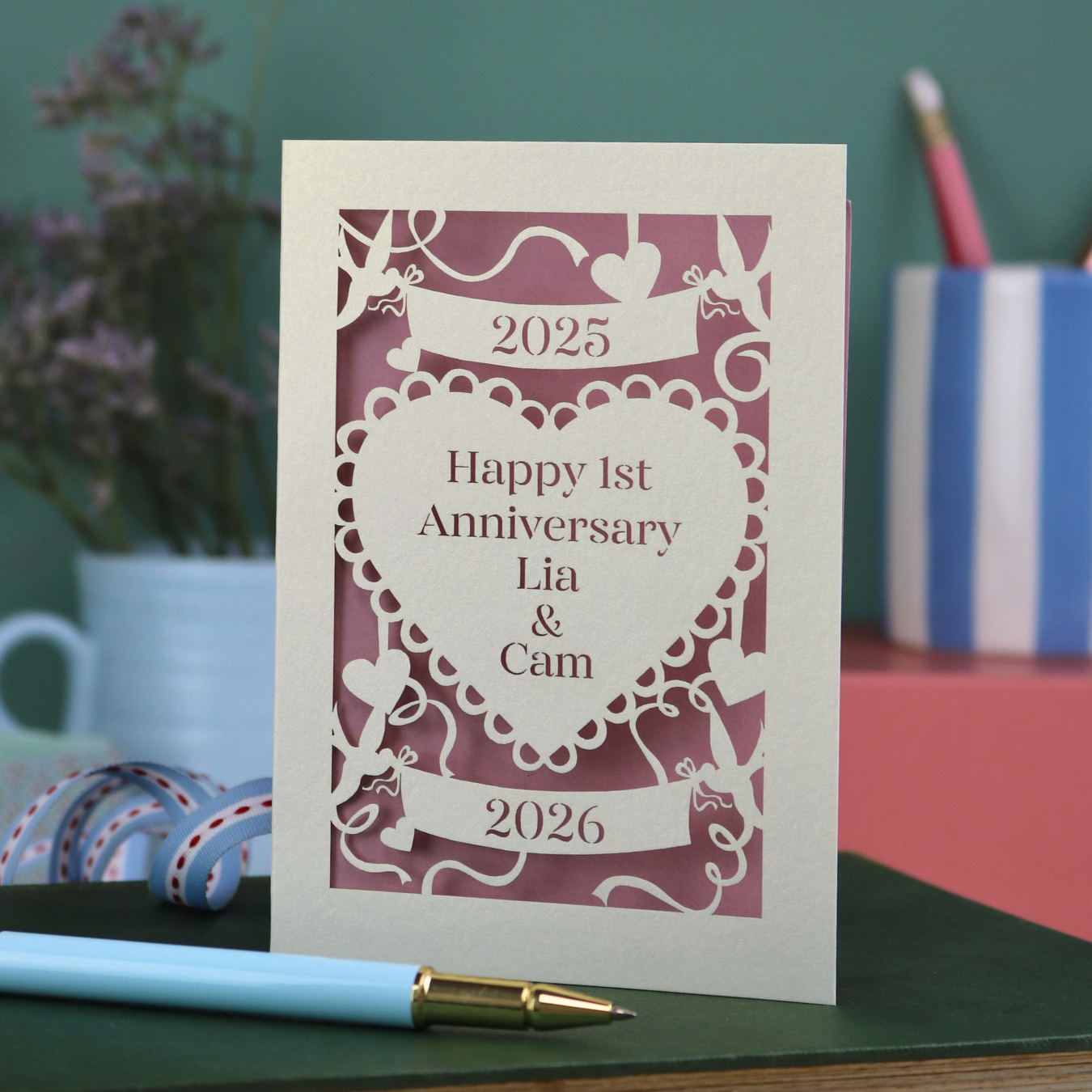 A laser cut cream first anniversary card with banners and heart design and the text "Happy 1st Anniversary Lia & Cam" with a banner above and the year 2025, and a banner below with the year 2026