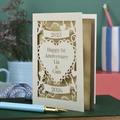 A laser cut cream first anniversary card with banners and heart design and the text "Happy 1st Anniversary Lia & Cam" with a banner above and the year 2025, and a banner below with the year 2026