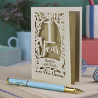 Decorative first anniversary card with '1 Year' and names, placed on a surface with a pen and blurred background.