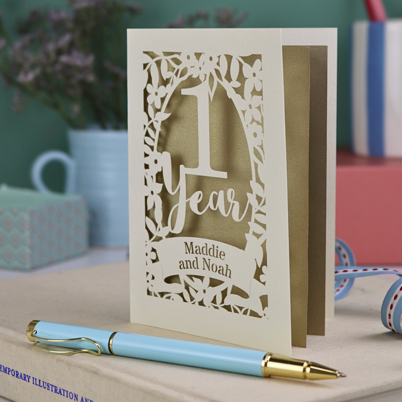 Decorative first anniversary card with '1 Year' and names, placed on a surface with a pen and blurred background.