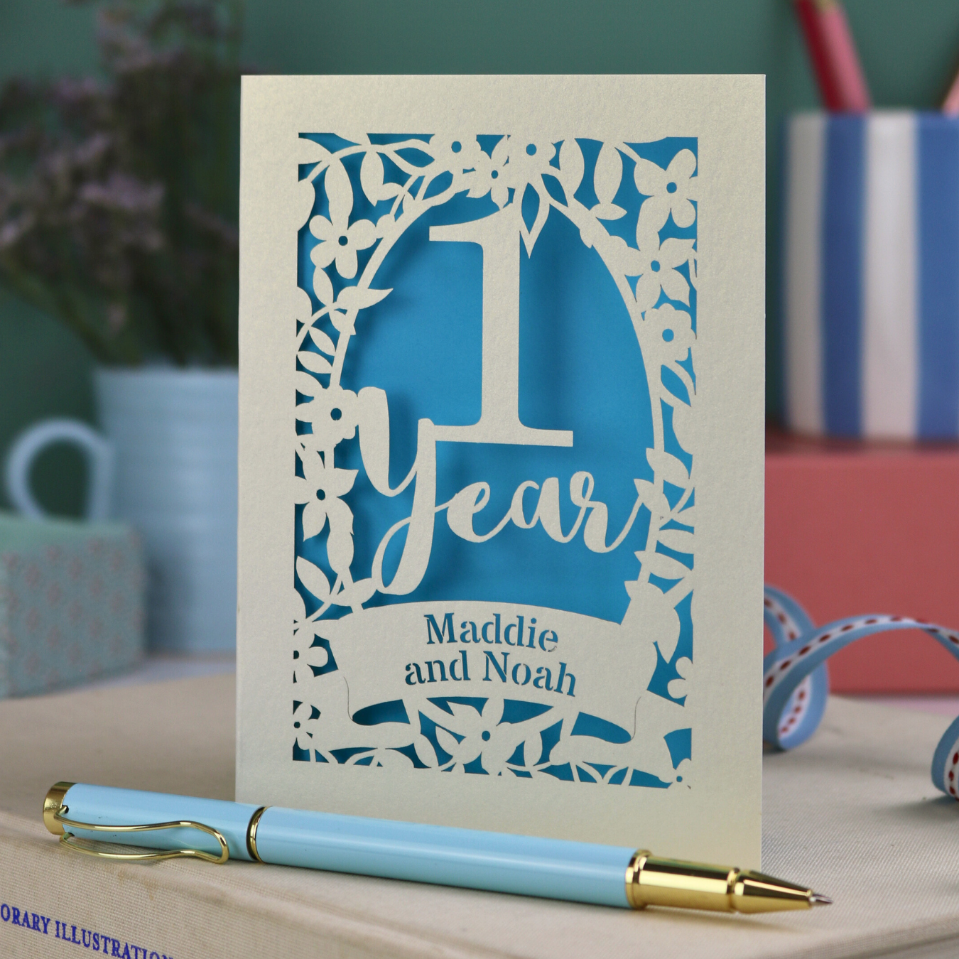 1st Wedding anniversary Card with '1 Year' and 'Maddie and Noah' text, accompanied by a blue pen on a desk.