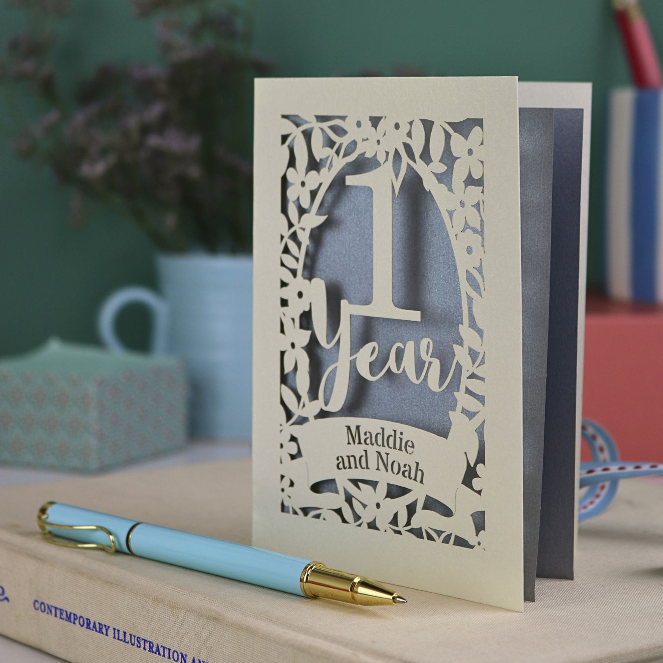 Personalised paper anniversary card with decorative cut-out design on a table with a pen and book.
