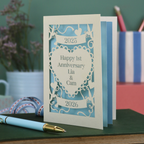 A laser cut  first anniversary card with banners and heart design and the text "Happy 1st Anniversary Lia & Cam" with a banner above and the year 2025, and a banner below with the year 2026