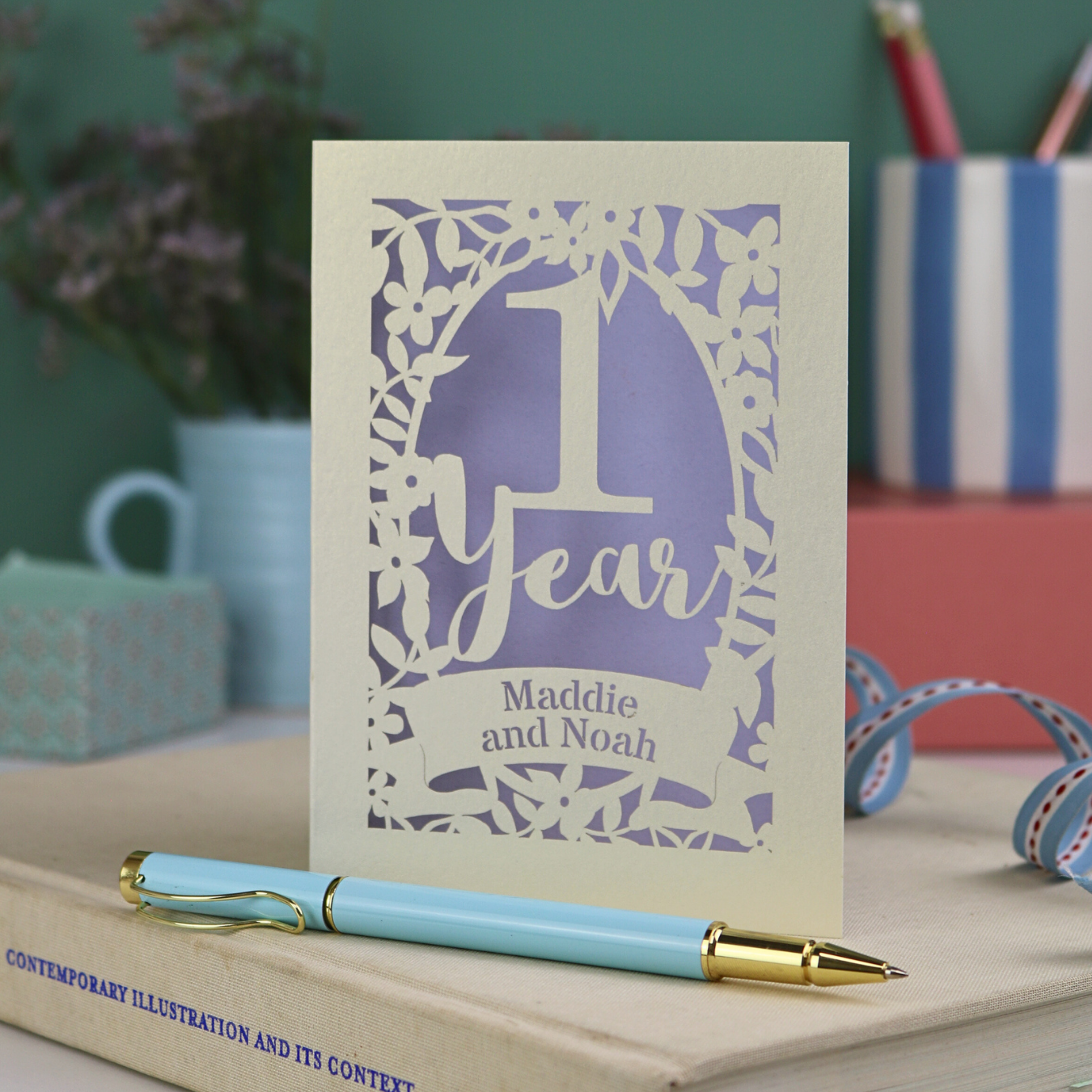 Laser cut first wedding Card with '1 Year' design on a book with a pen next to it, against a blurred background.