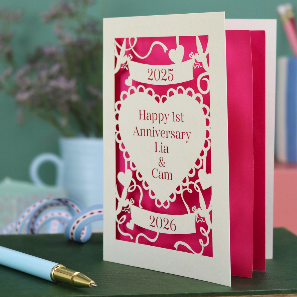 A laser cut cream first anniversary card with banners and heart design and the text "Happy 1st Anniversary Lia & Cam" with a banner above and the year 2025, and a banner below with the year 2026