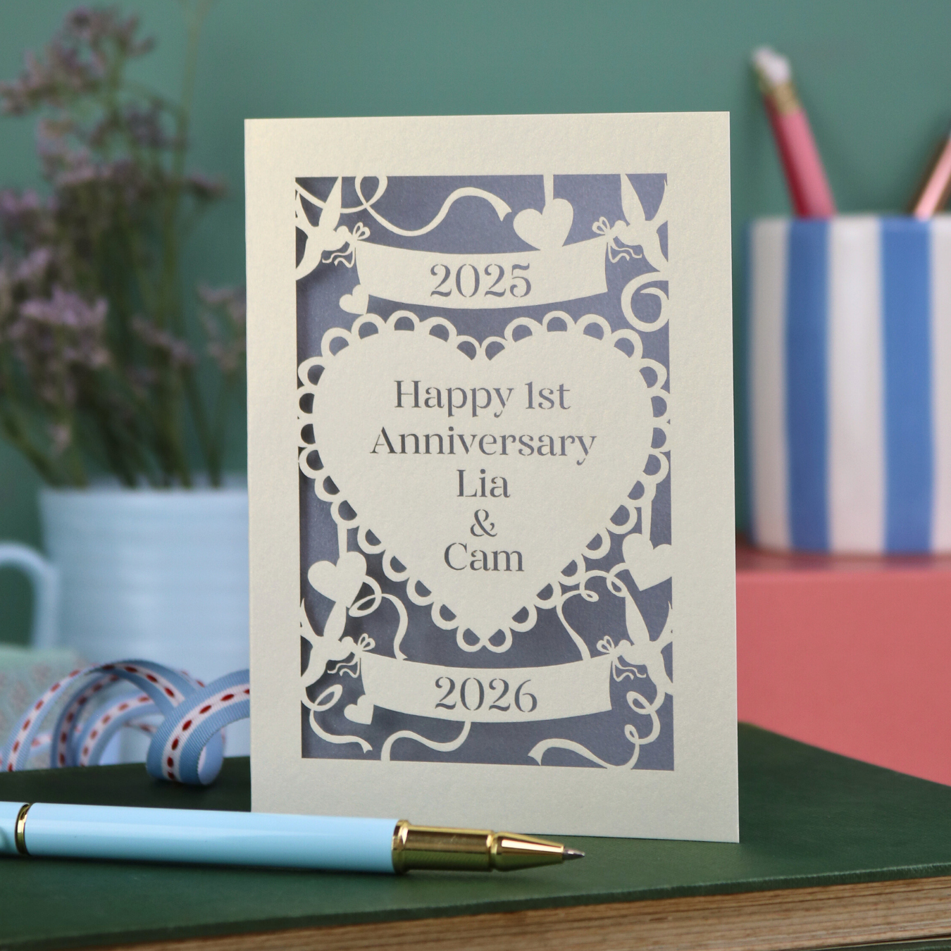 A laser cut cream first anniversary card with banners and heart design and the text "Happy 1st Anniversary Lia & Cam" with a banner above and the year 2025, and a banner below with the year 2026