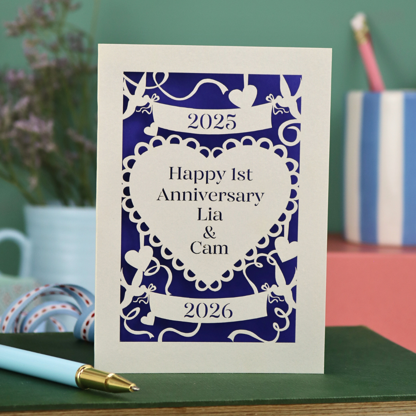 A laser cut cream 1st anniversary card with banners and heart design and the text "Happy 1st Anniversary Lia & Cam" with a banner above and the year 2025, and a banner below with the year 2026