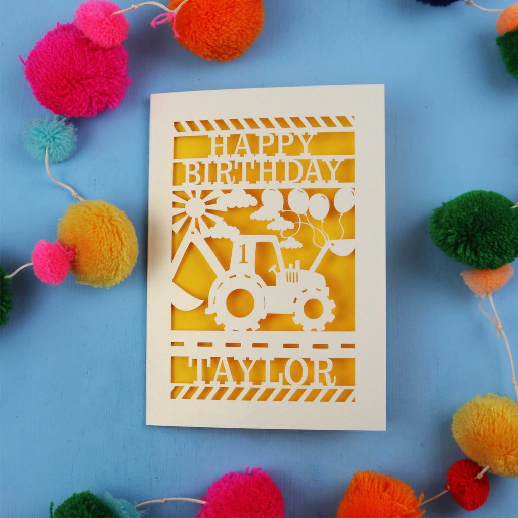 1st Birthday card with a laser cut digger design and 'Happy Birthday Taylor' text on a blue background with colourful pom-poms.