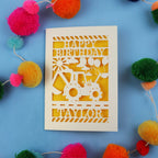 1st Birthday card with a laser cut digger design and 'Happy Birthday Taylor' text on a blue background with colourful pom-poms.