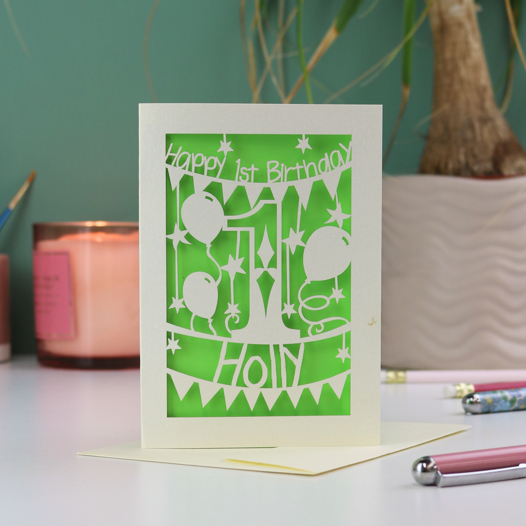 Green birthday card with cream cut-out designs on a table with a candle and pen.