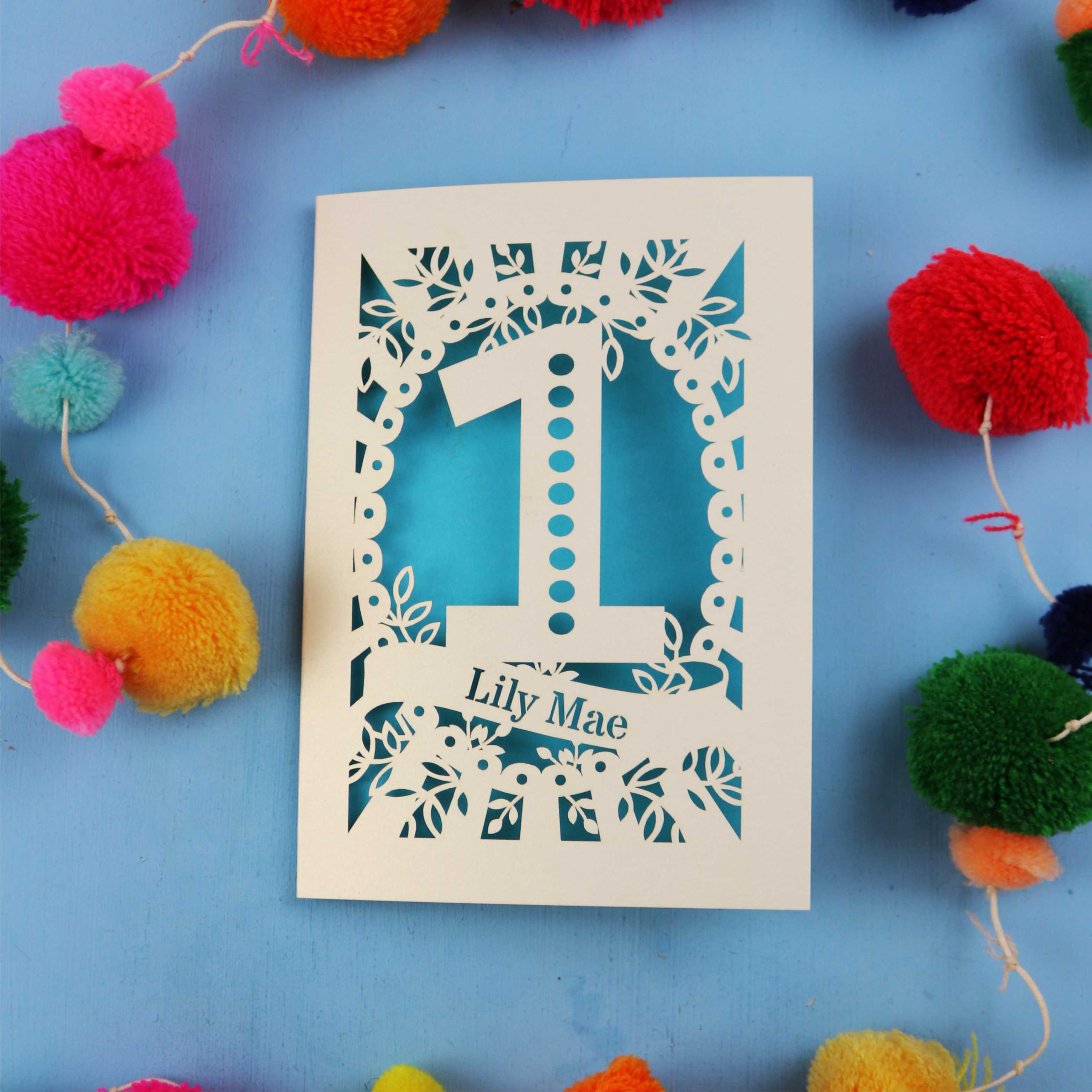 Decorative First birthday card with '1' and 'Lily Mae' on a blue background with colourful pom-poms.