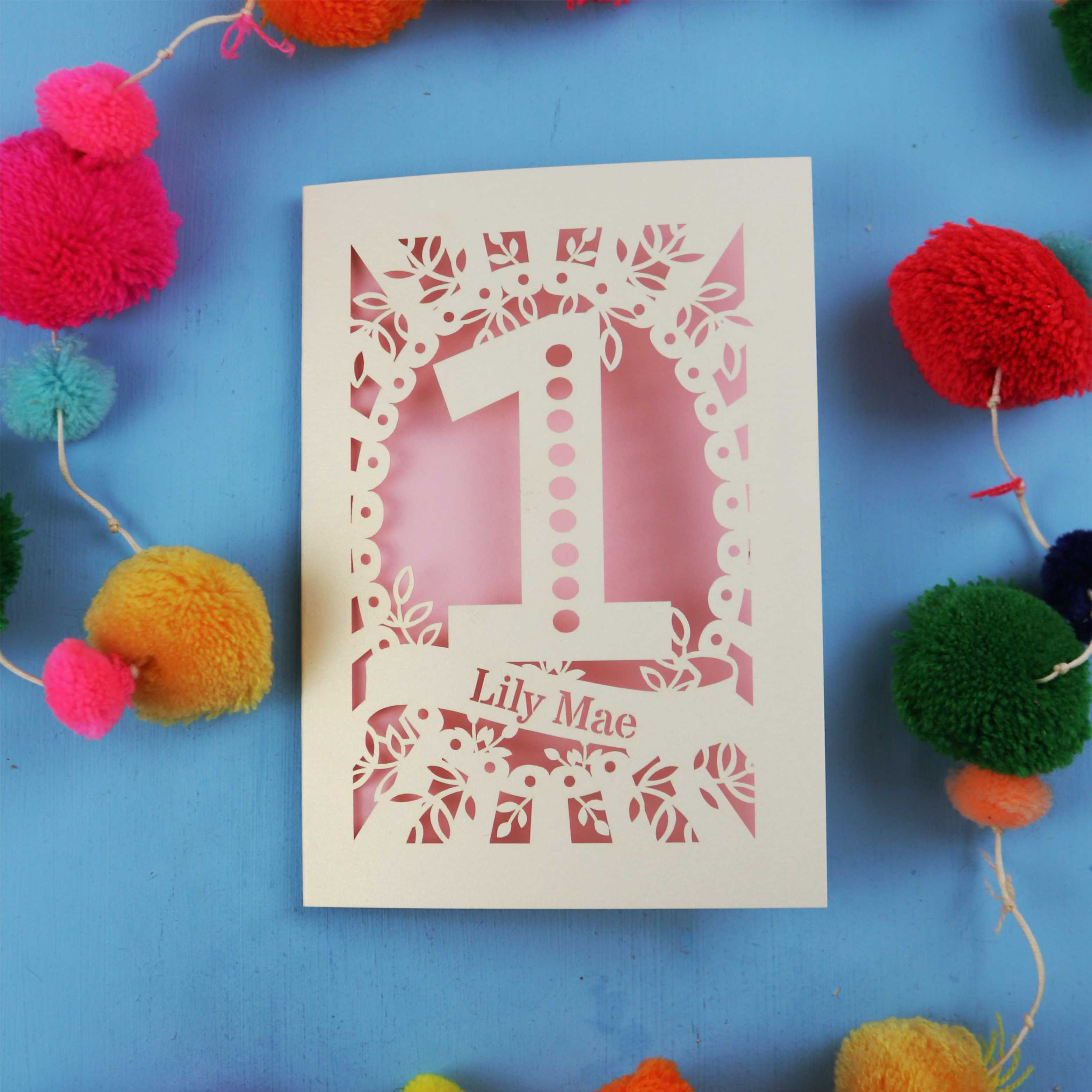 Laser-cut 1st birthday card with number '1' and name 'Lily Mae' on a blue background with colourful pom-poms.