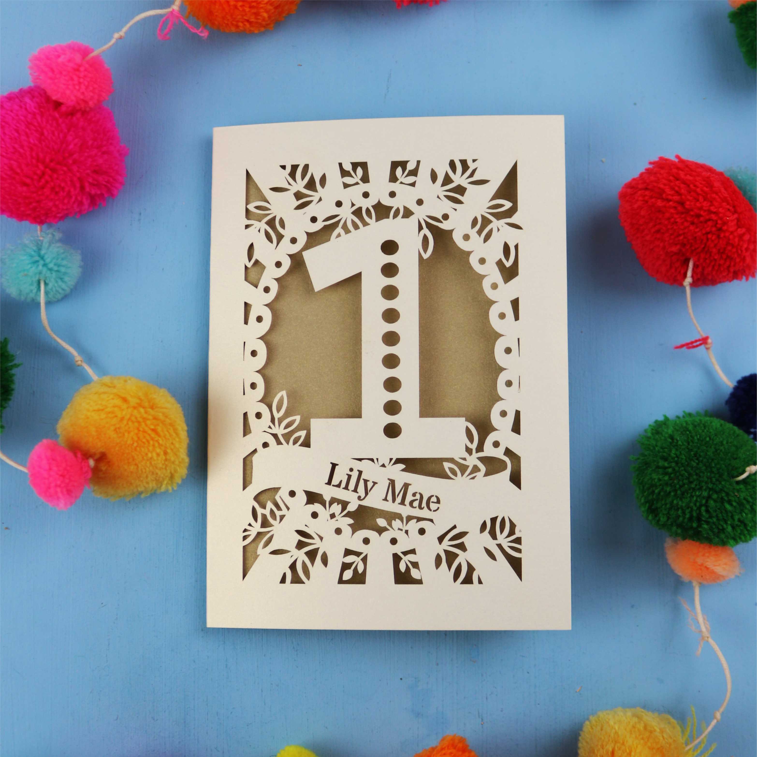 Laser-cut birthday card with number '1' and name 'Lily Mae' on a blue background with colourful pom-poms.