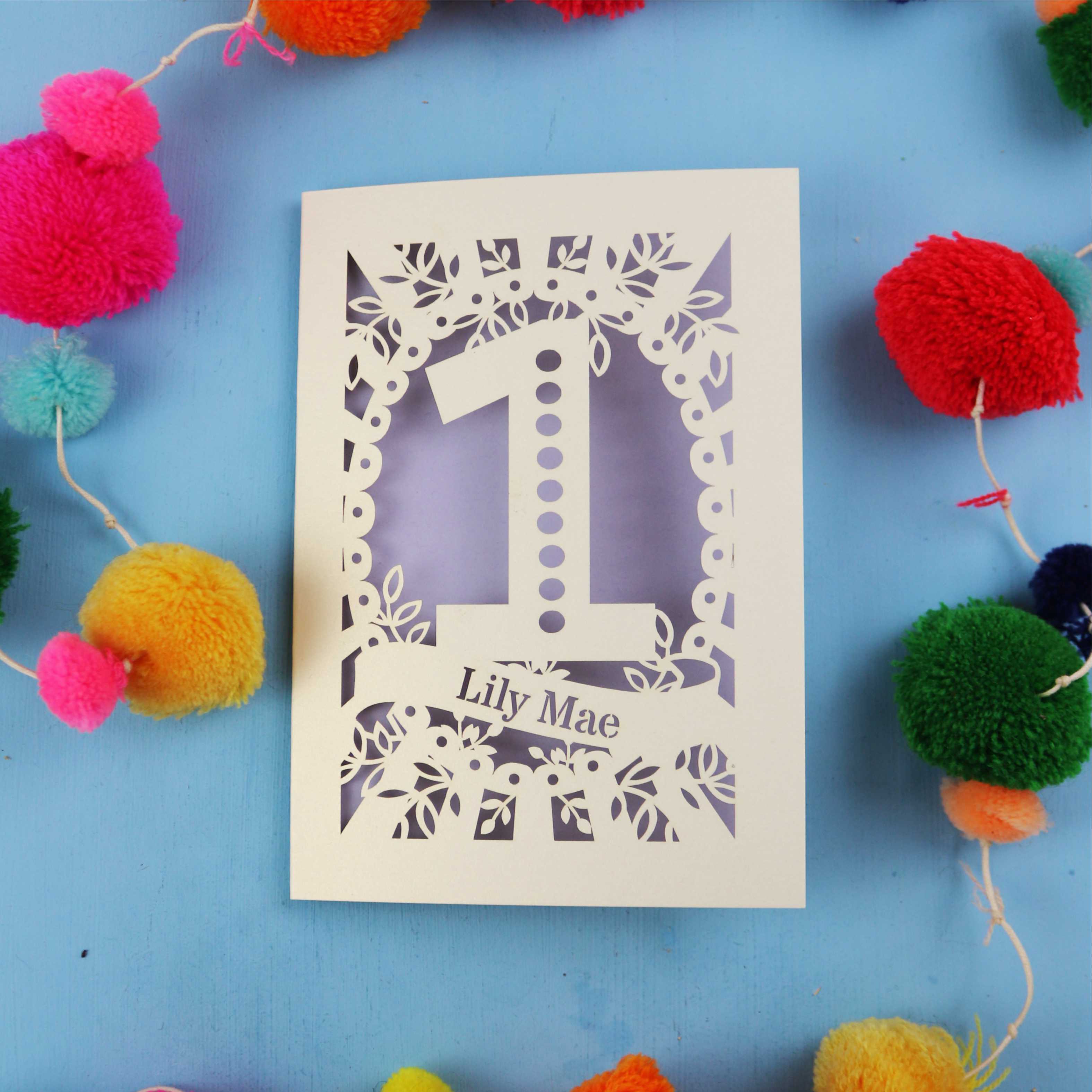 Laser-cut card with '1' and 'Lily Mae' text on a blue background with colourful pom-poms.