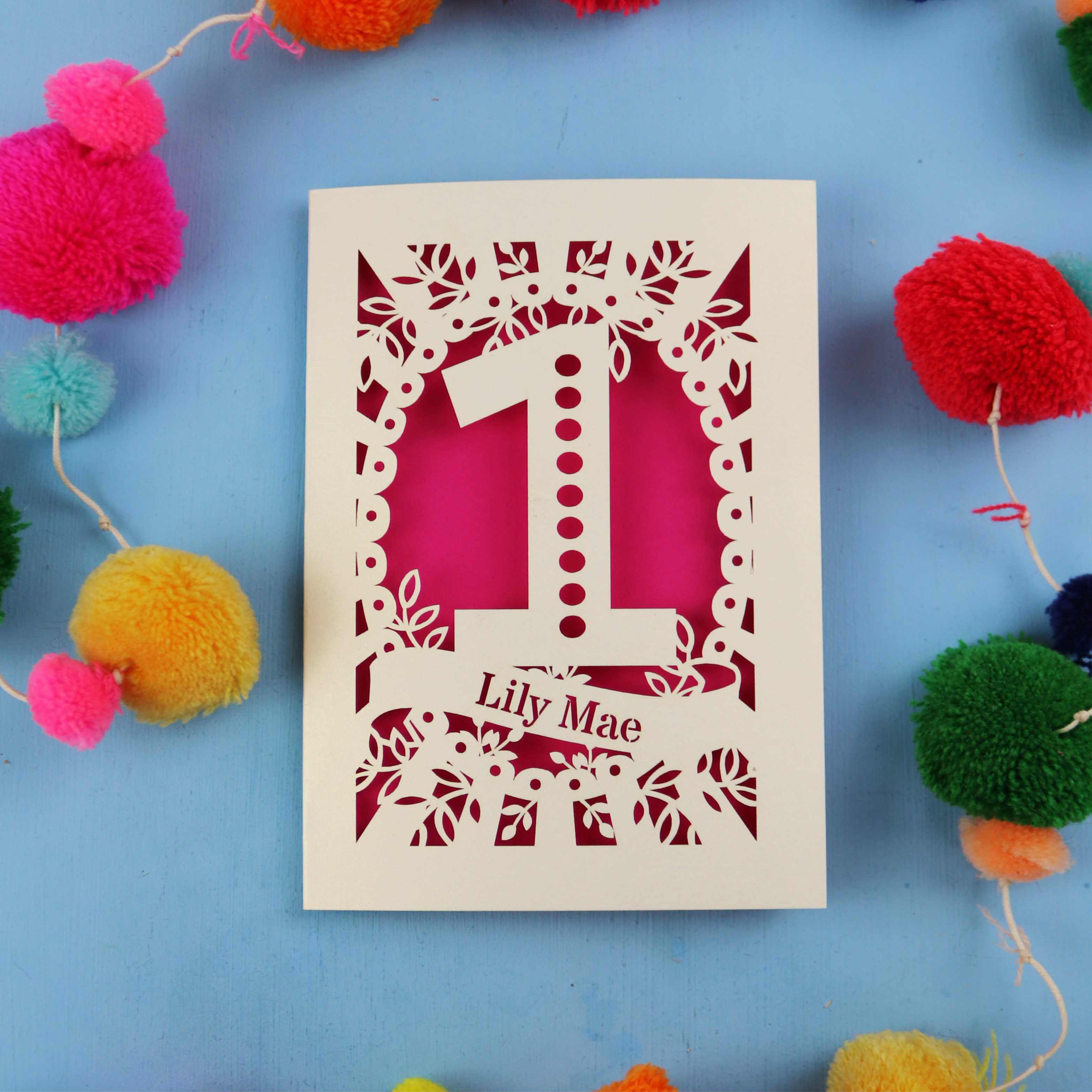 Laser-cut first birthday card with number 1 and name 'Lily Mae' on a blue background with colourful pom-poms.