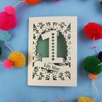 Laser-cut 1 Birthday card with number '1' and name 'Lily Mae' on a blue background with colourful pom-poms.