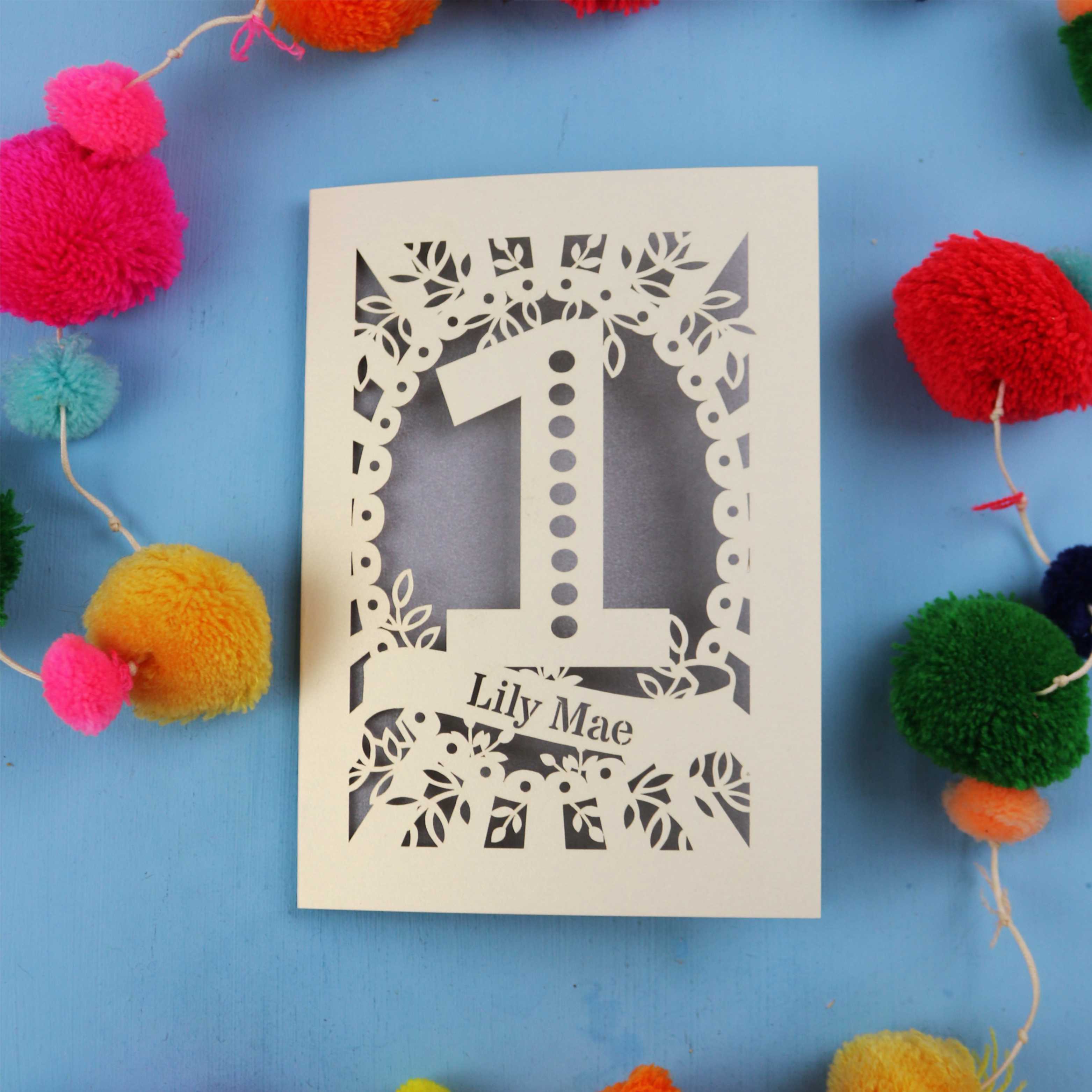 Laser-cut card with number '1' and name 'Lily Mae' on a blue background with colourful pom-poms.
