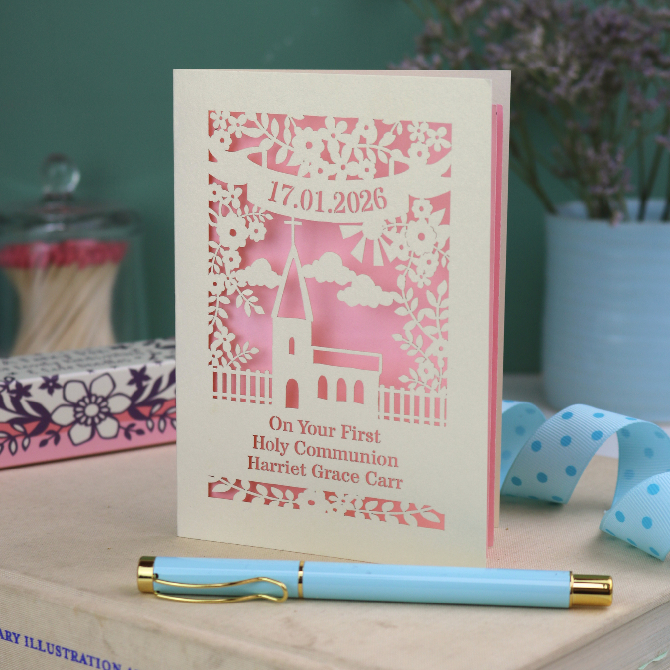 A cream and candy pink laser cut 'On Your First Holy Communion' card with cut out church design, floral border, the date and name in cut out stencil font.