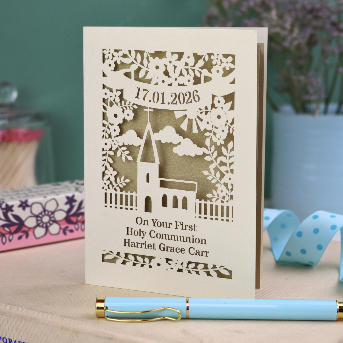 Personalised card with laser-cut design of a church and text, "On Your First Holy Communion Harriet Grace Carr" and the date, on a wooden surface with a pen and decorative items.