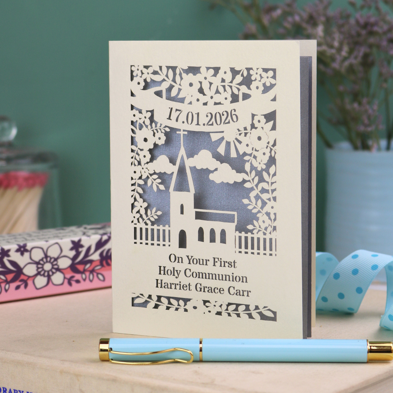 A cream and silver paper cut 'On Your First Holy Communion' card with cut out church design, floral border, personalised with the date and name in cut out stencil font.