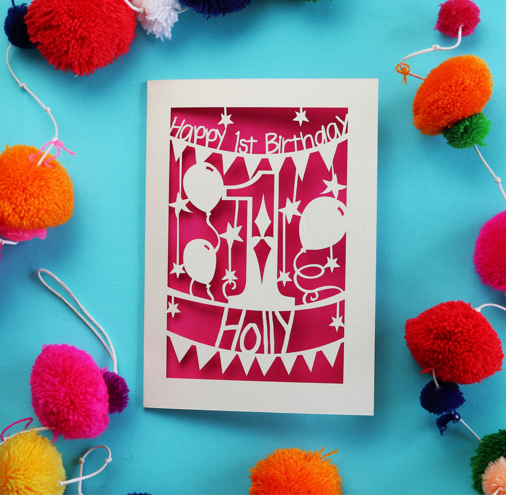 A personalised paper cut birthday card. Card says Happy 1st Birthday, with a big number 1 and balloons in the middle. Card is personalised with a name underneath and bunting. Card is cream and shocking pink.