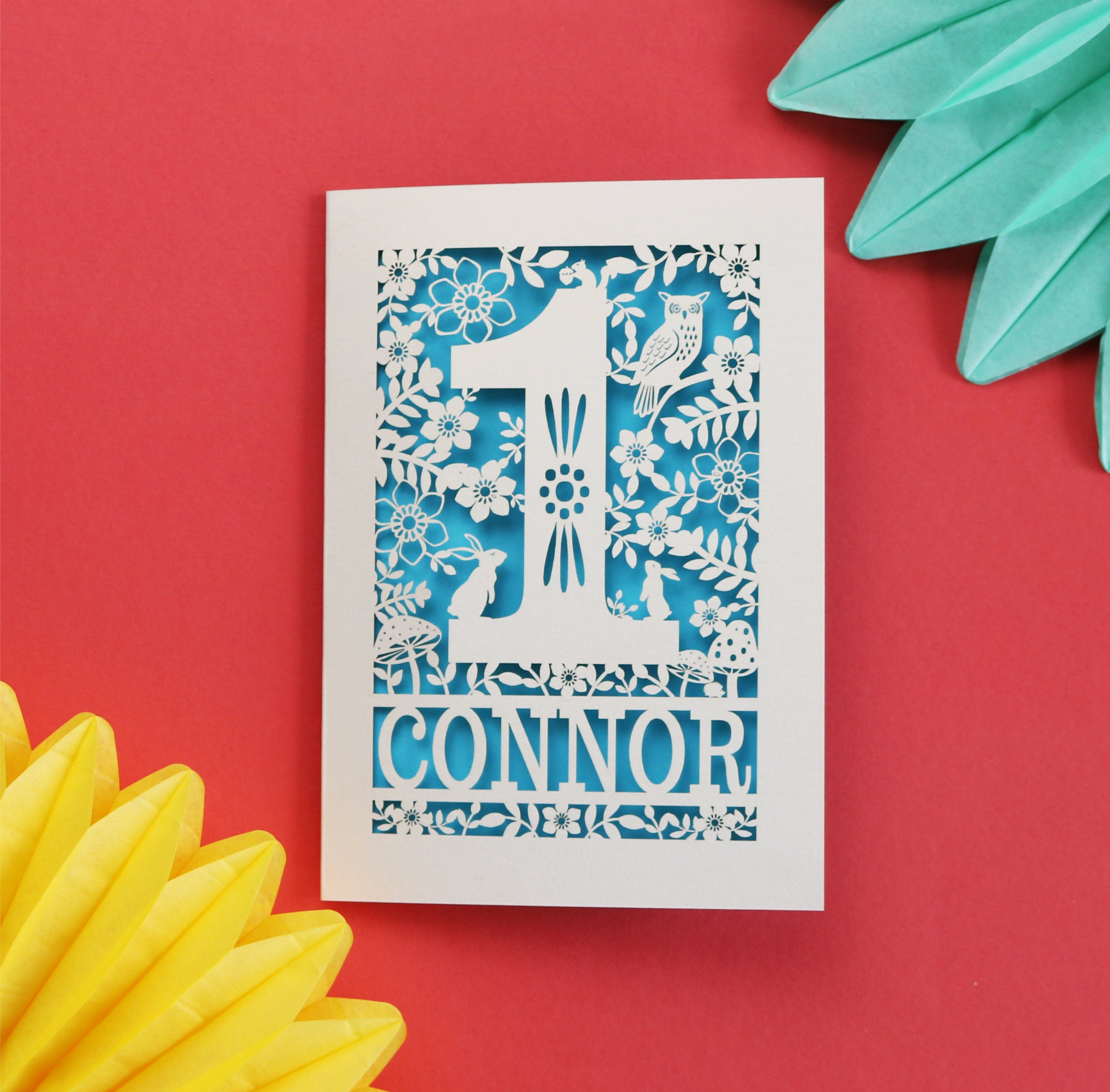 A personalised papercut 1st birthday card with a woodland animal theme, featuring cut-out shapes of an owl, rabbits, and a hedgehog, and the name 'CONNOR' in bold letters.