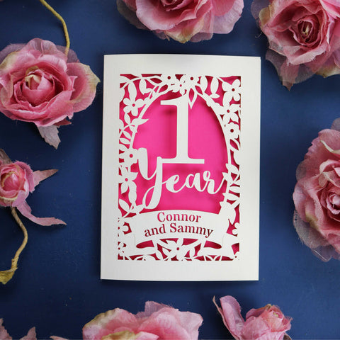 A cream and shocking pink paper cut wedding anniversary card. Card has "1 Year" written in calligraphy font and surrounded by a floral border. Names are cut out of a banner.  - A5 (large); / Shocking Pink;