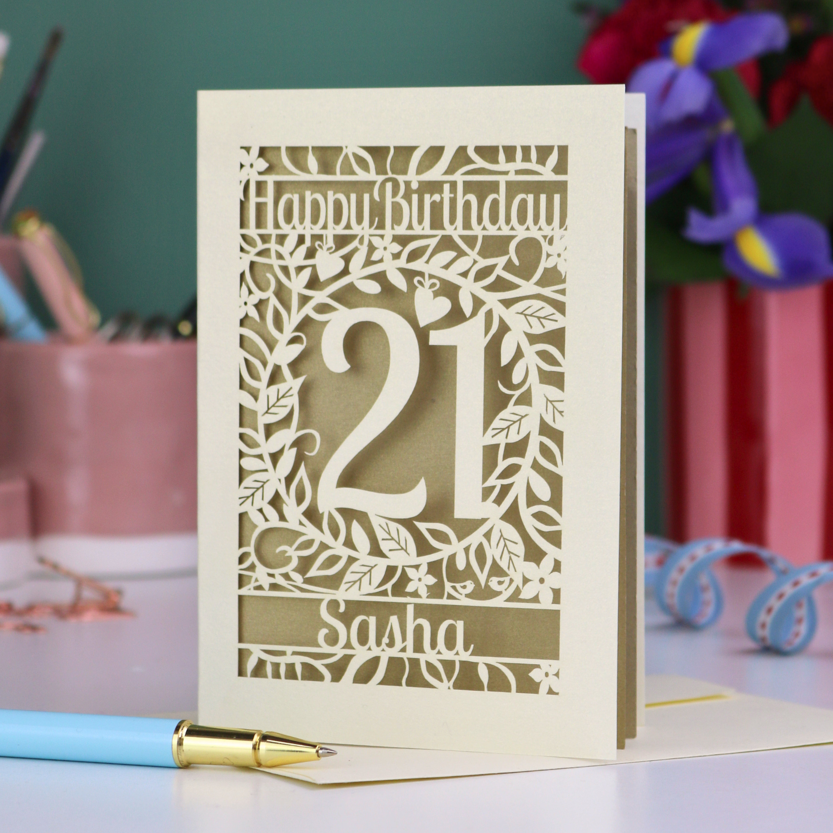 Personalised 21st Birthday card with 'Happy Birthday Sasha' and laser cut floral design on a desk with stationery items.