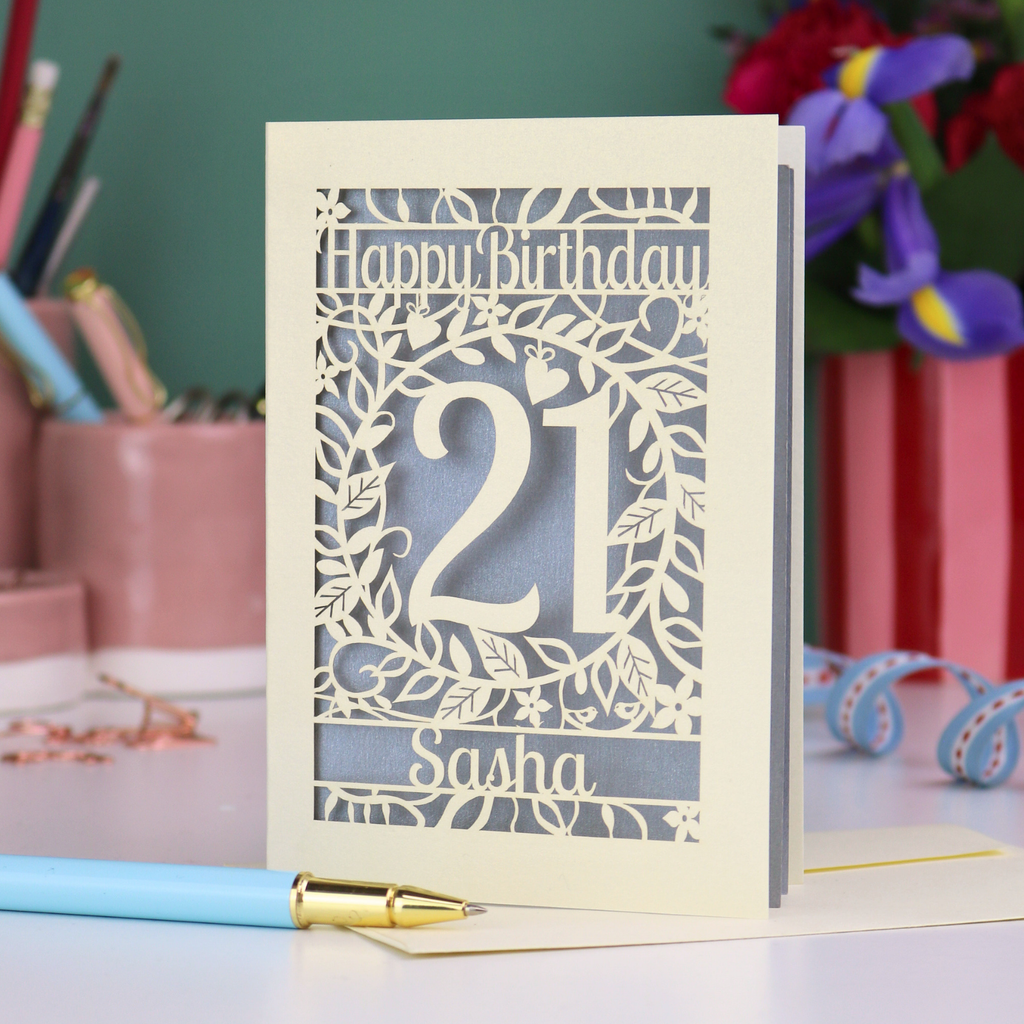 Personalised 21st Birthday card with 'Happy Birthday Sasha' and laser cut floral design on a desk with stationery items.
