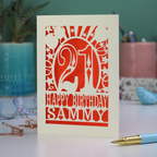 A laser cut 21st birthday card with 'Happy Birthday Sammy' in cut out text and the number 21 in a bold circus font, with balloons, stars and sunbeams border,  on a desk with stationery items.