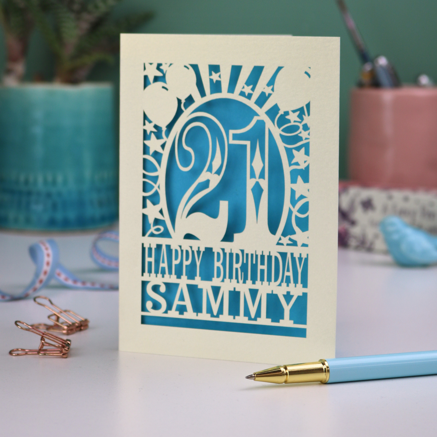 A laser cut 21st birthday card with 'Happy Birthday Sammy' in cut out text and the number 21 in a bold circus font, with balloons, stars and sunbeams border,  on a desk with stationery items.