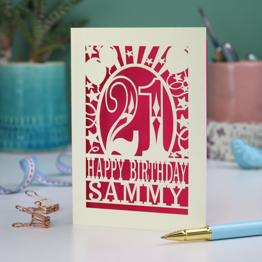 A laser cut 21st birthday card with 'Happy Birthday Sammy' in cut out text and the number 21 in a bold circus font, with balloons, stars and sunbeams border,  on a desk with stationery items.