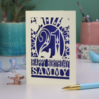 A laser cut 21st birthday card with 'Happy Birthday Sammy' in cut out text and the number 21 in a bold circus font, with balloons, stars and sunbeams border,  on a desk with stationery items.