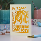 A laser cut 21st birthday card with 'Happy Birthday Sammy' in cut out text and the number 21 in a bold circus font, with balloons, stars and sunbeams border,  on a desk with stationery items.