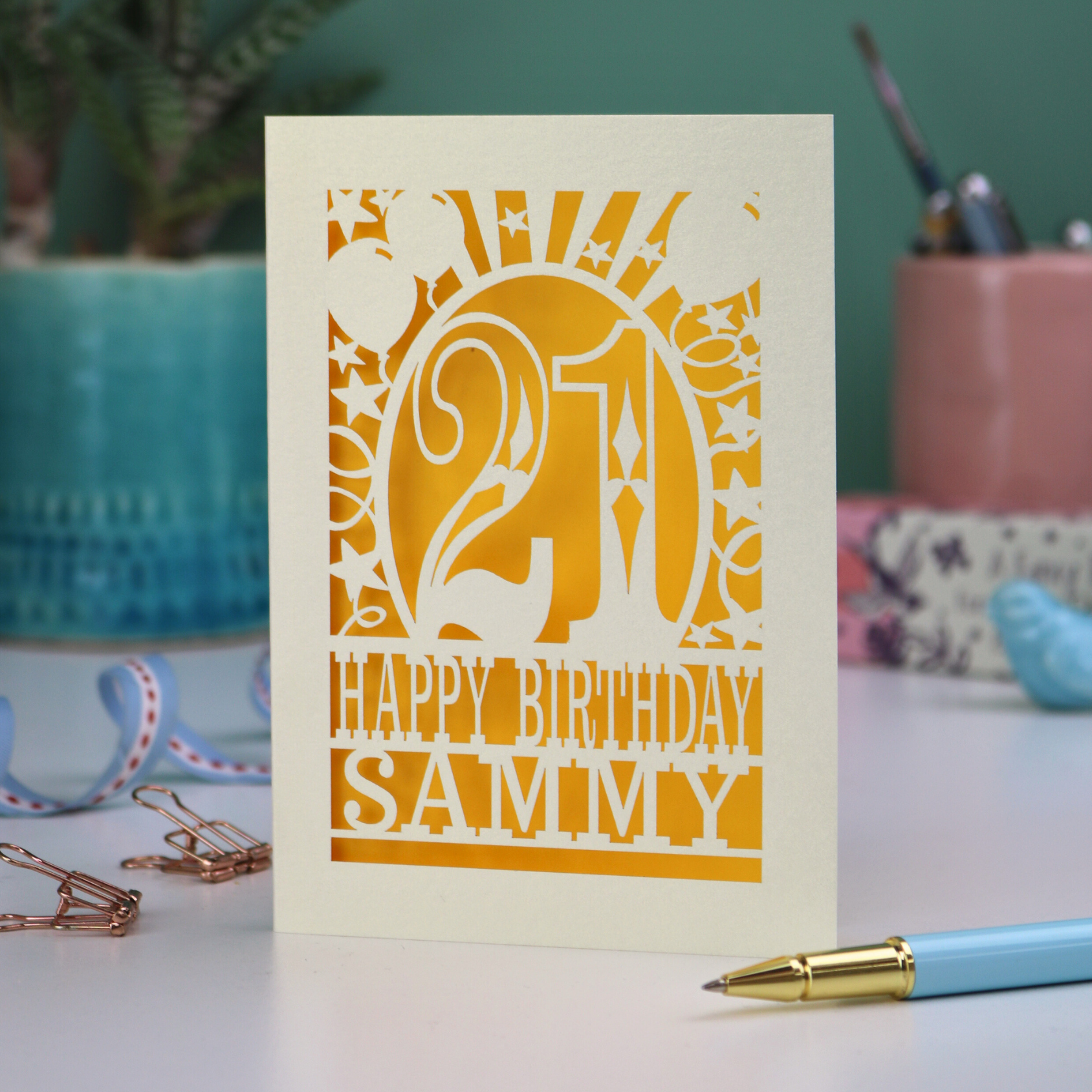 A laser cut 21st birthday card with 'Happy Birthday Sammy' in cut out text and the number 21 in a bold circus font, with balloons, stars and sunbeams border,  on a desk with stationery items.