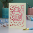 Decorative laser cut 21st birthday card with cupcake design and 'Georgie' text on a table with stationery items.