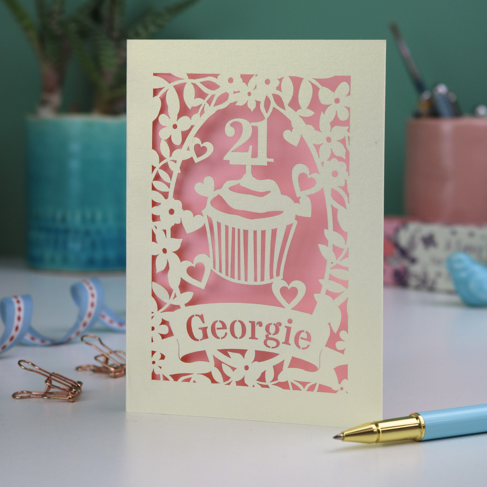 Decorative laser cut 21st birthday card with cupcake design and 'Georgie' text on a table with stationery items.