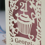 Decorative laser cut 21st birthday card with cupcake design and 'Georgie' text on a table with stationery items.