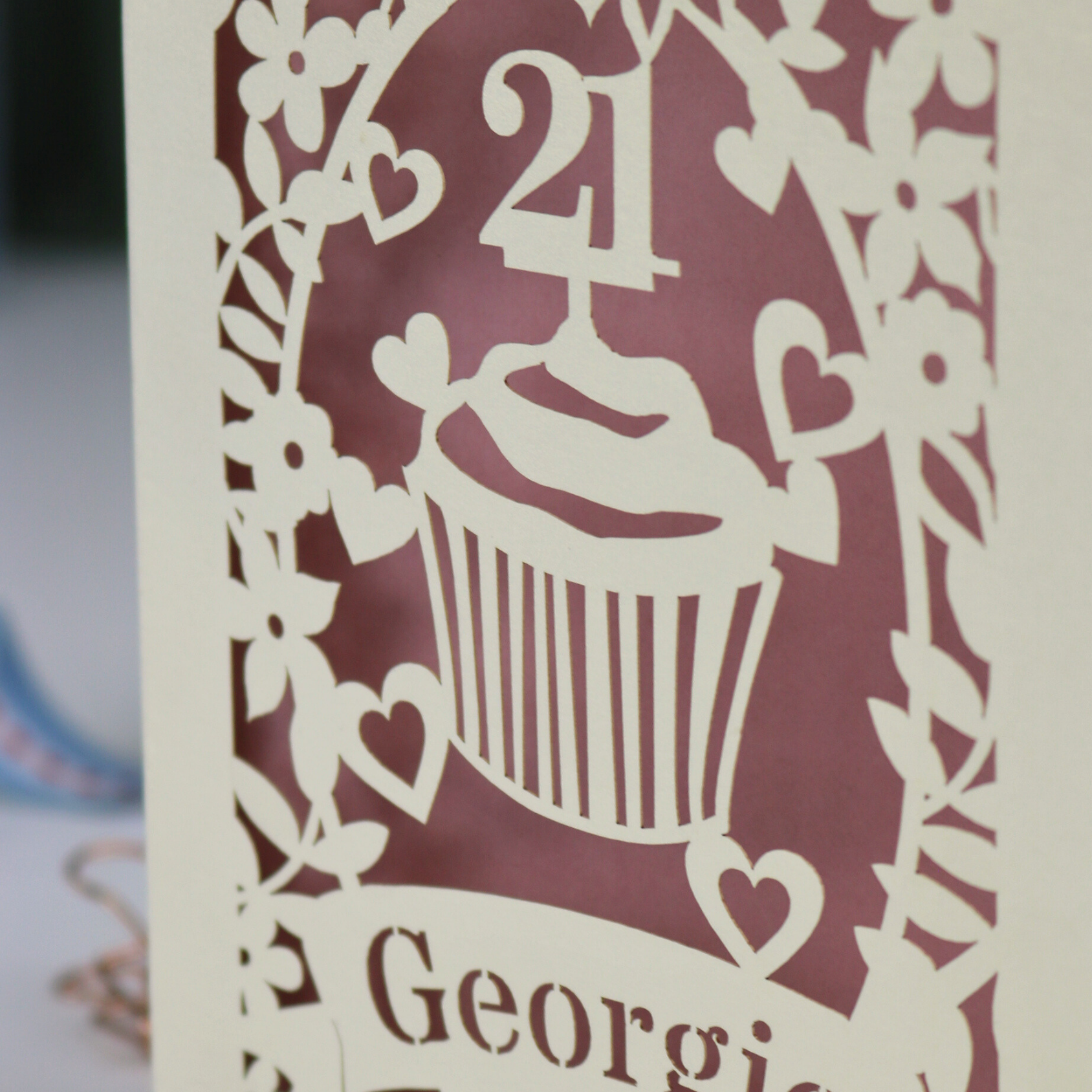 Decorative laser cut 21st birthday card with cupcake design and 'Georgie' text on a table with stationery items.